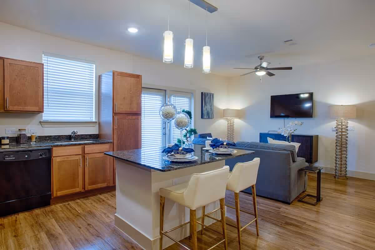 Open concept interior space featuring a kitchen with wooden cabinets, a black countertop island with two white bar stools, and a living area with a gray sofa, wall-mounted TV, two modern floor lamps, and a ceiling fan. The room has wooden flooring and large windows with blinds.