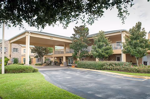 Exterior view of a two-story senior living facility with a covered entrance driveway, surrounded by green lawns, bushes, and trees under a partly cloudy sky.