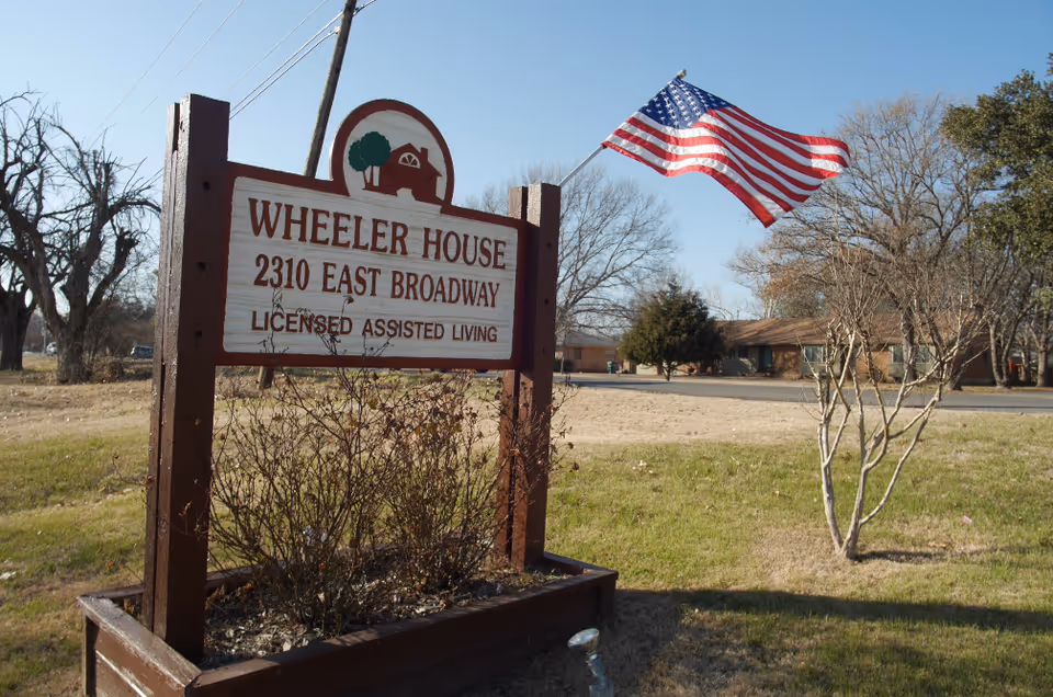 Wooden sign for Wheeler House reading '2310 East Broadway Licensed Assisted Living' with an American flag and a single-story building behind it.