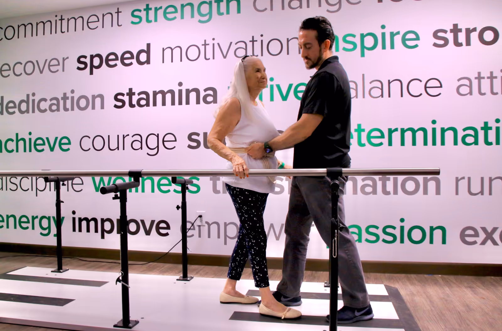 An elderly woman is assisted by a male therapist while walking on a parallel bar setup in a rehabilitation or physical therapy room. The wall behind them is decorated with motivational words such as strength, speed, stamina, determination, and passion in various fonts and colors.
