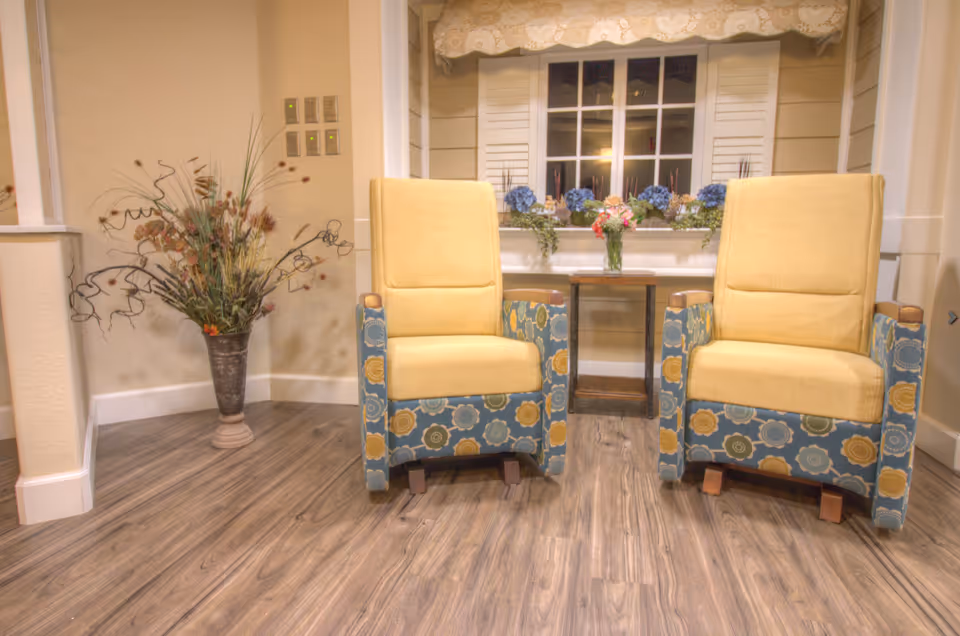 Two upholstered armchairs with yellow cushions and floral patterned sides are placed on a wooden floor in front of a window with white shutters and a flower box filled with blue and pink flowers. A small wooden side table with a vase of flowers sits between the chairs. To the left, there is a tall decorative vase with dried plants and a beige wall with light switches.