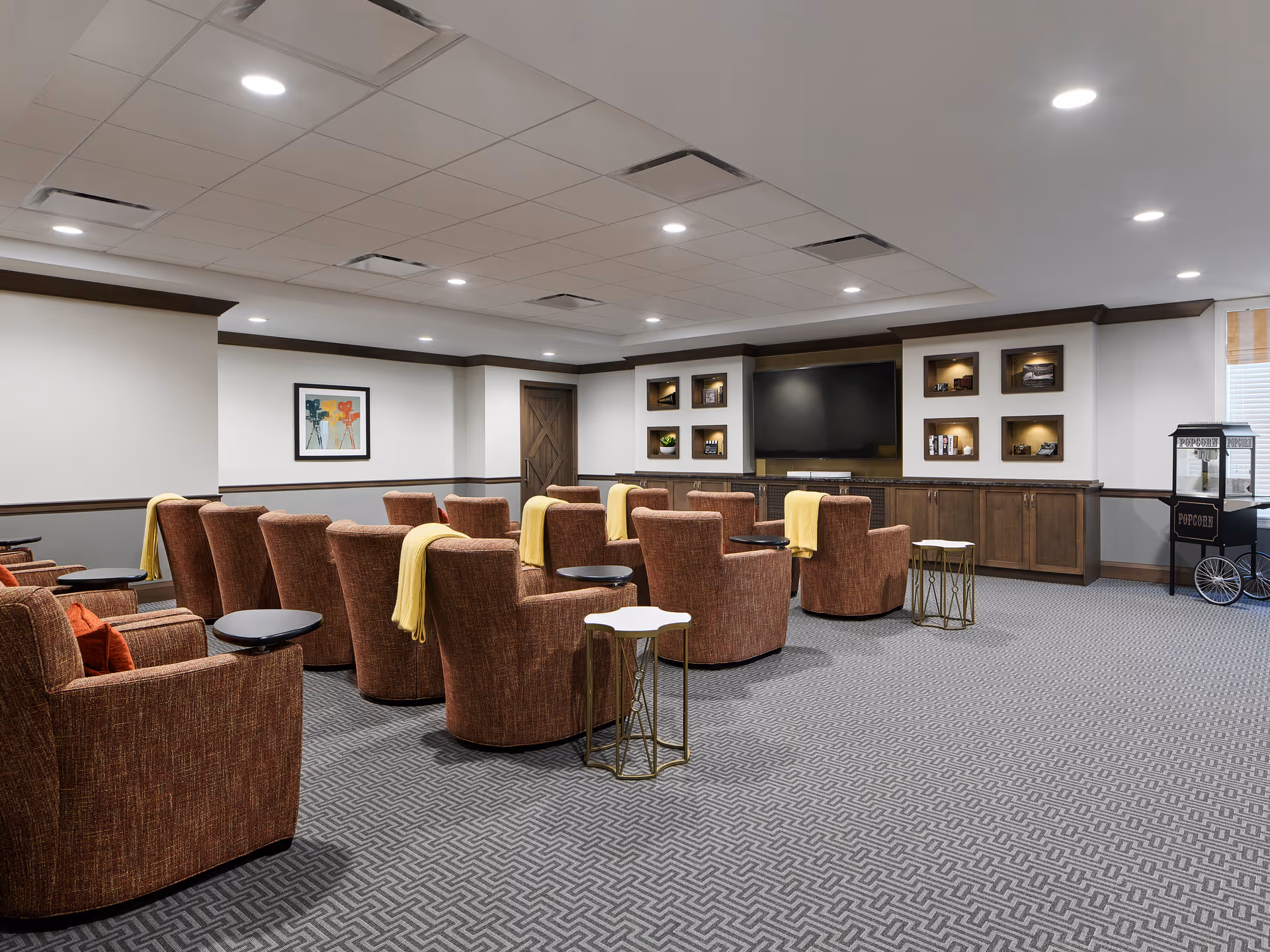 A cozy media room with multiple brown upholstered armchairs arranged in rows facing a large flat-screen TV mounted on a wall with built-in shelves displaying decorative items. Small side tables are placed beside some chairs, and yellow throws are draped over the backs of several chairs. The room has a patterned carpet, recessed ceiling lights, and a popcorn machine in the corner near a window with blinds.
