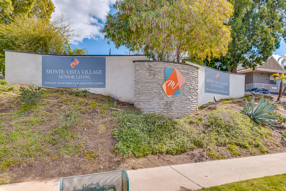 Entrance sign for Monte Vista Village Senior Living on a landscaped stone wall with trees and a sidewalk in front.