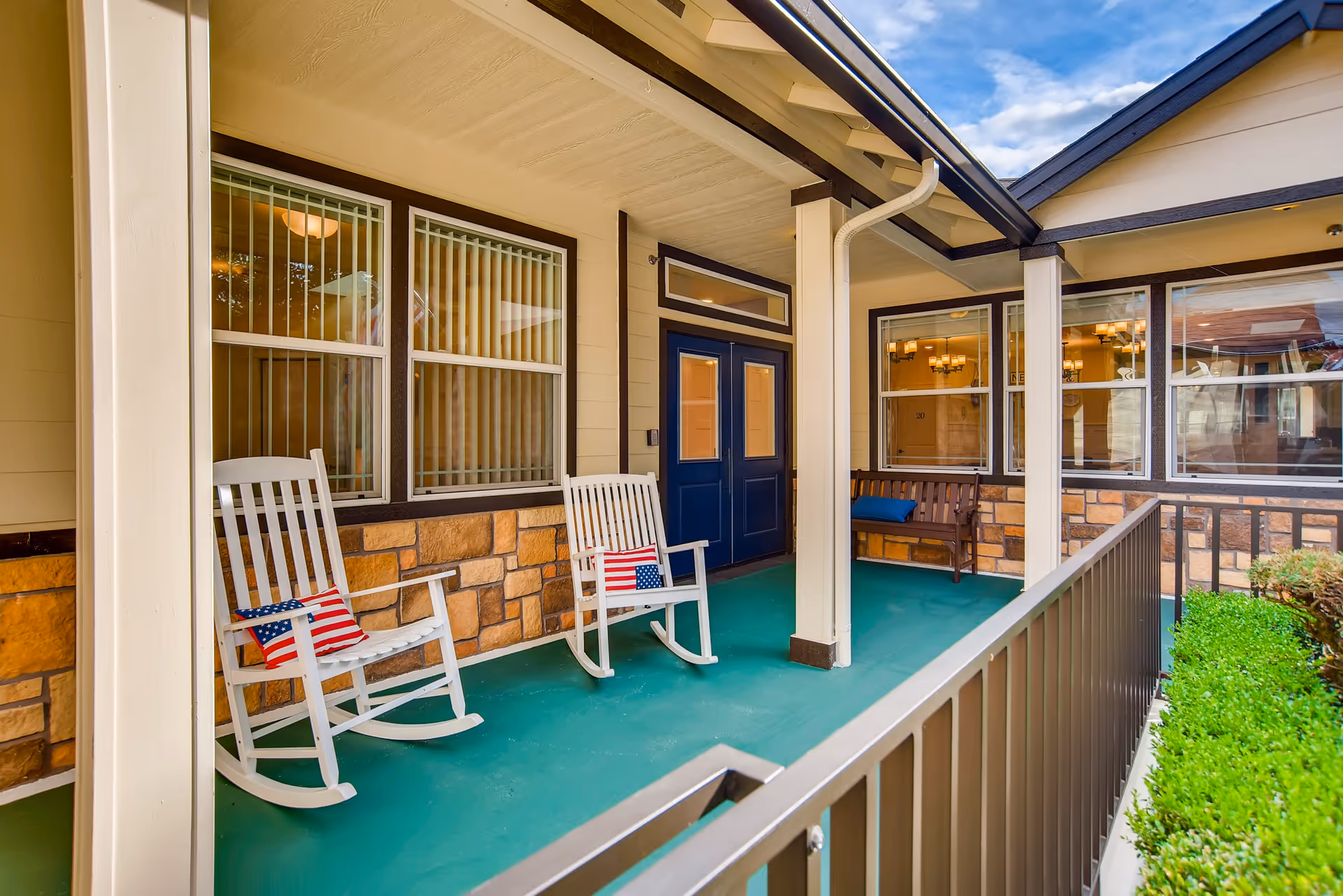 Covered porch area with two white rocking chairs each with a small American flag pillow, a wooden bench with a blue cushion, stone and beige siding walls, blue double doors, and windows with vertical blinds. The porch has a green floor and is bordered by a metal railing and some greenery.