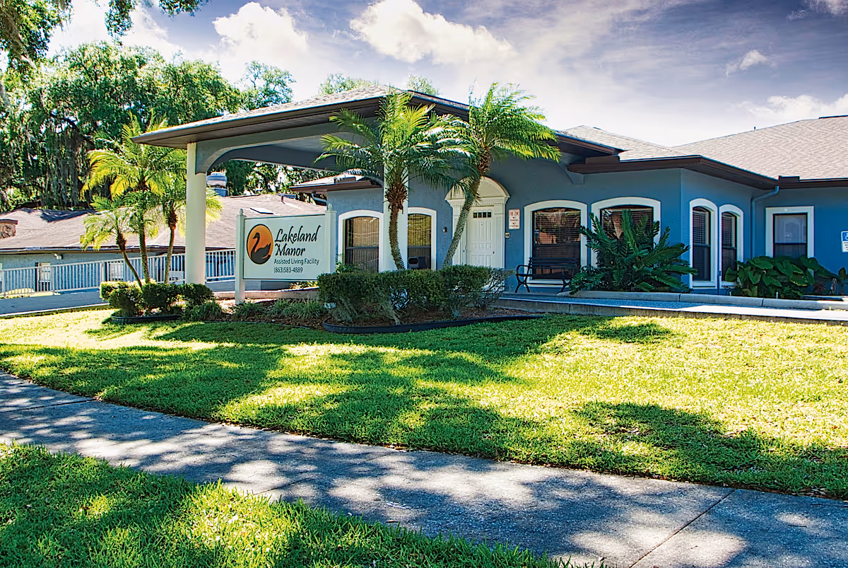 Single-story blue assisted living facility front with a covered entrance, sign reading 'Lakeland Manor Assisted Living Facility', palm trees and a grassy lawn.