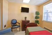 A small, simple room with a single bed covered with a red and green blanket, a wooden chair with blue upholstery, a dark wooden dresser with a small plant on top, a flat-screen TV mounted on the wall, and a tall green plant near a window with blinds.
