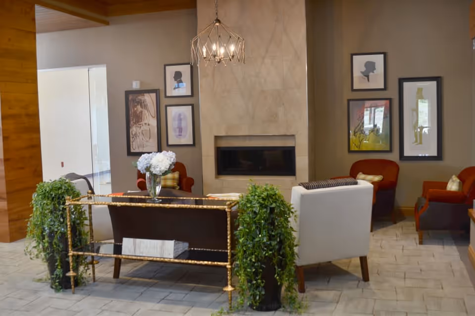 A cozy living room area with a modern fireplace centered on a beige wall. The room features a dark brown sofa with a glass vase of white flowers on a table behind it, a white armchair, and two red armchairs. Green plants flank the sofa, and framed abstract art hangs on the walls. A geometric chandelier hangs from the ceiling.