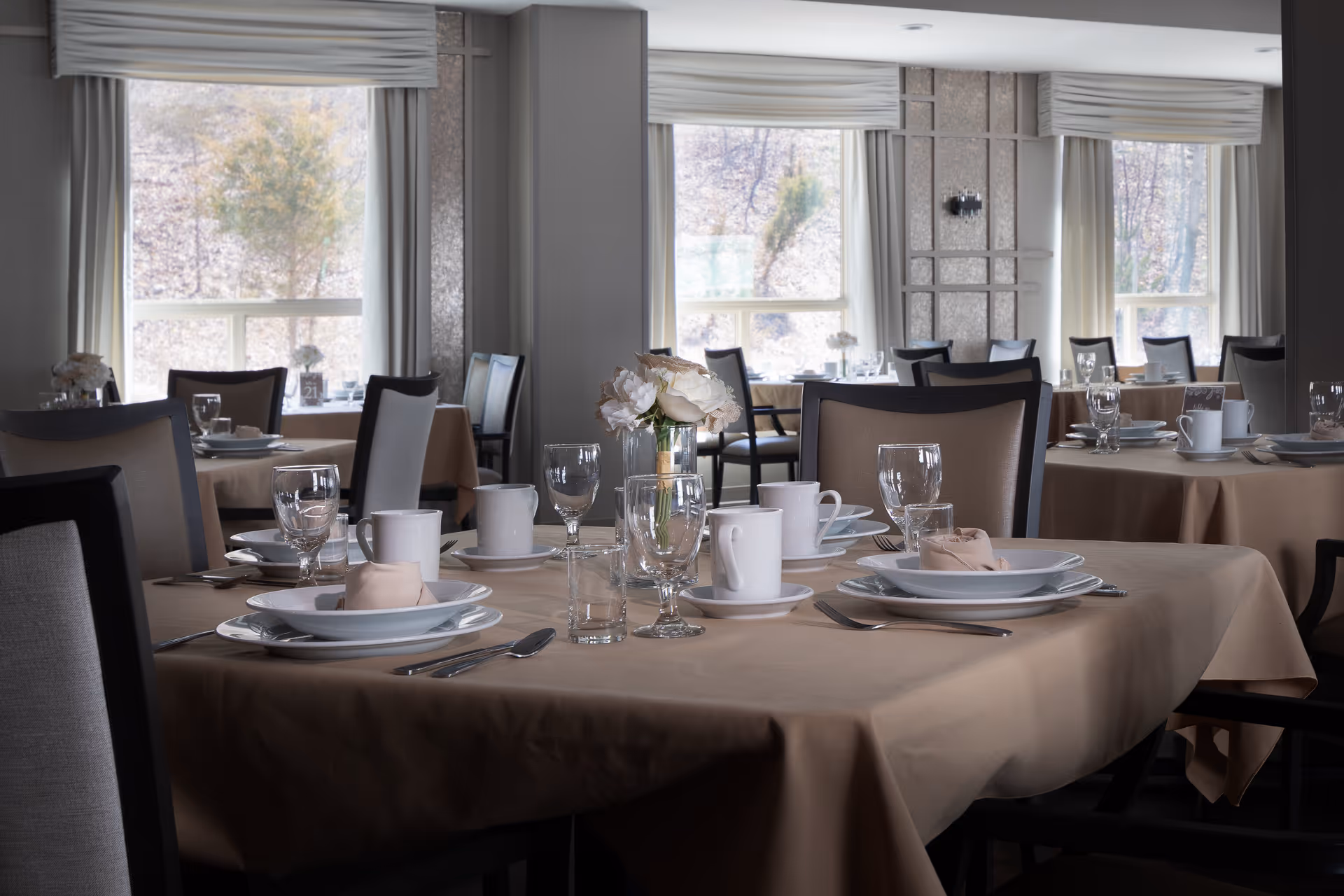 A dining room with multiple tables set for a meal, featuring beige tablecloths, white plates, cups, glassware, silverware, and floral centerpieces. Large windows with light curtains allow natural light to brighten the room.