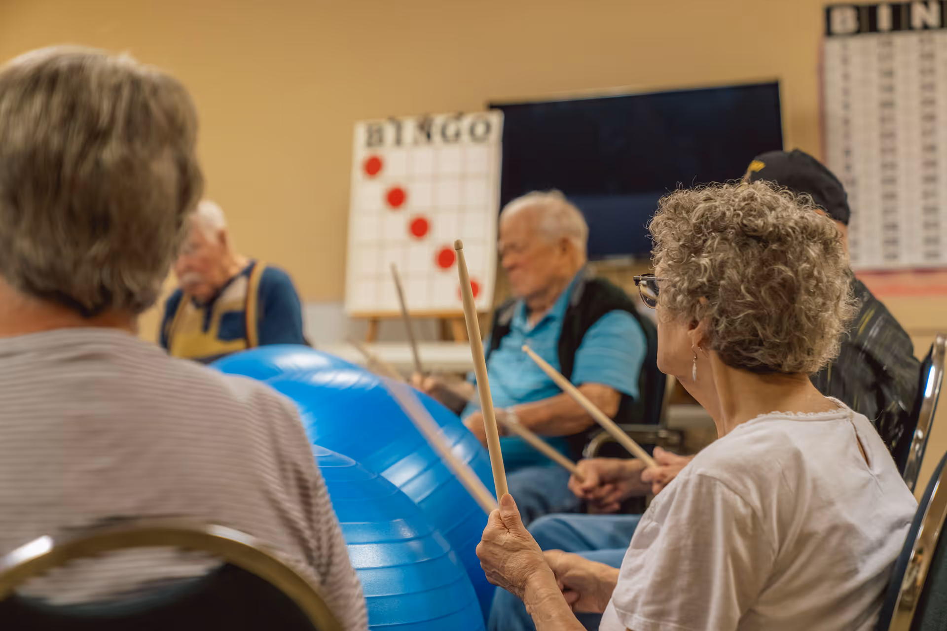 A group of elderly people sitting in chairs in a room, participating in a drumming activity using drumsticks and large blue exercise balls. In the background, there is a bingo board with red markers and a television screen.