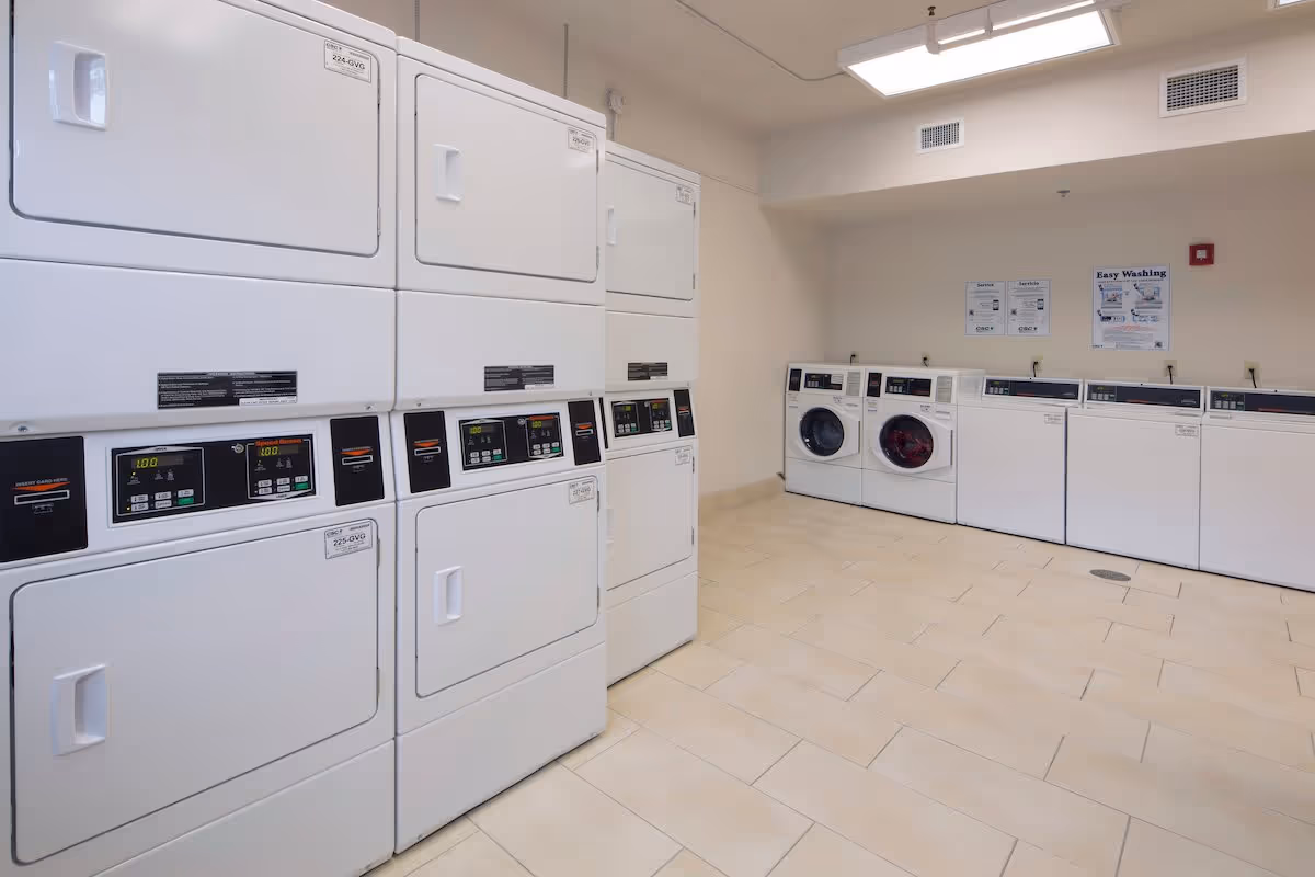 A clean laundry room with multiple white washing machines and dryers. The machines are stacked on the left side and lined up along the back wall. The room has beige tiled flooring and white walls with instructional posters on the back wall.