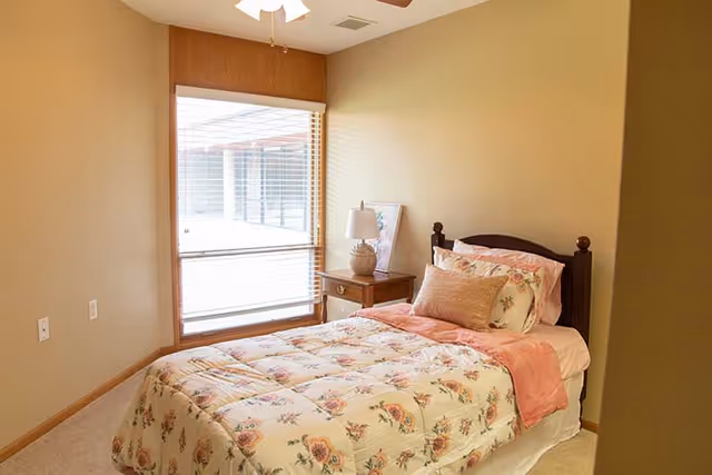 Small bedroom with a single bed dressed in floral bedding, a wooden nightstand with a lamp, and a large window with blinds.