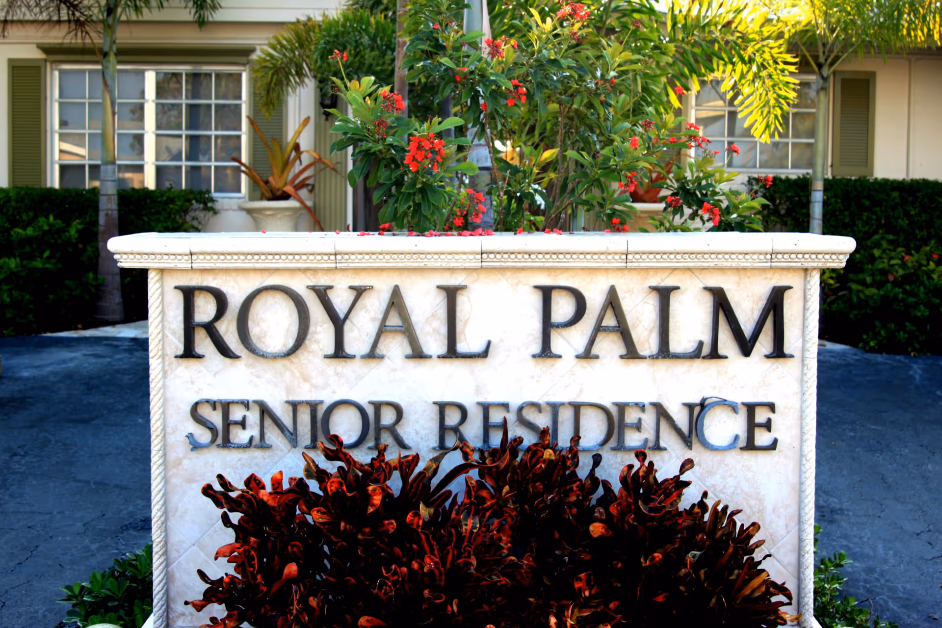 Stone sign reads Royal Palm Senior Residence with surrounding tropical plants and a building in background.
