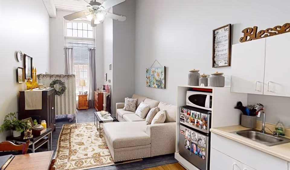 Open-plan living area with a beige sectional sofa, patterned rug, kitchenette with microwave and mini-fridge, and a tall window at the far end.
