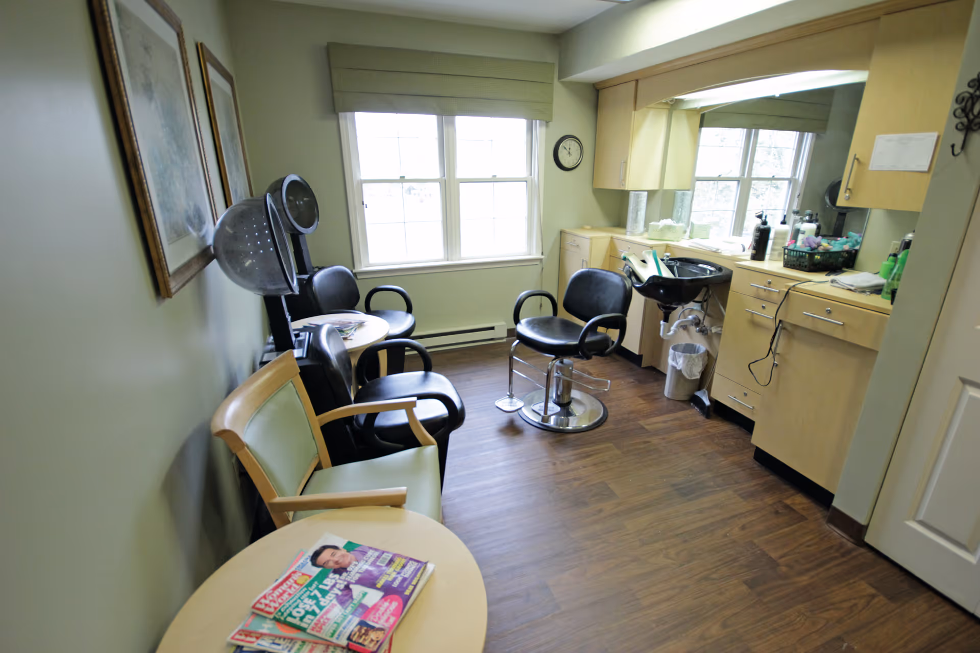 A small hair salon room with two black salon chairs, a hair washing sink, wooden cabinets, a window with a green valance, and a small round table with magazines. There is also a green cushioned chair and a hair dryer attached to the wall.