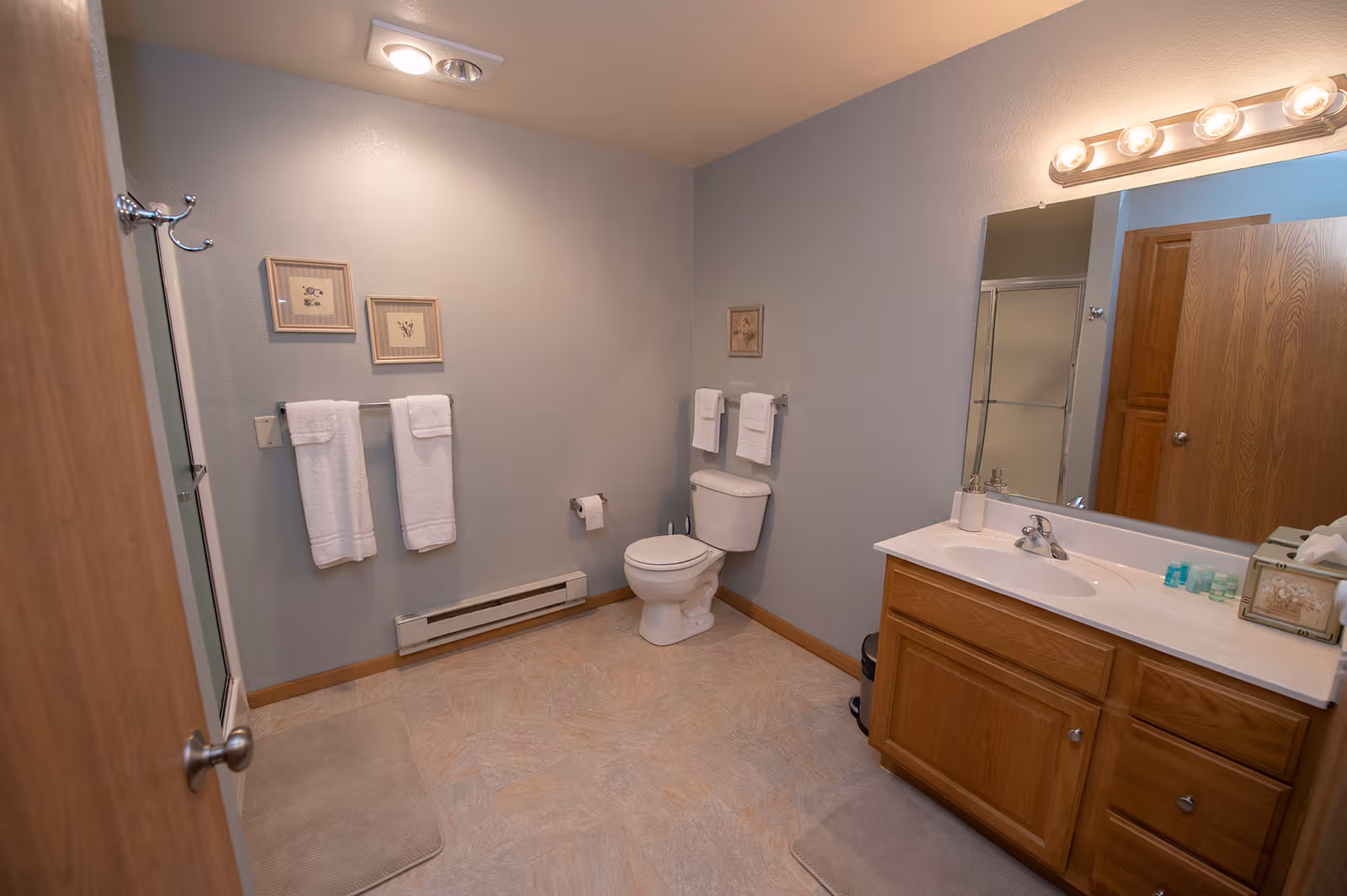 Spacious residential bathroom with a toilet, wooden vanity and sink with mirror, shower, towel racks and framed wall art.