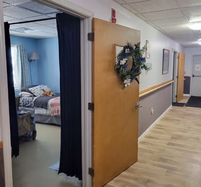 View of a hallway in a senior living facility named Wayne's Place, showing an open wooden door decorated with a floral wreath leading into a bedroom with a bed, pillows, and a stuffed animal. The hallway has wood flooring, handrails on the wall, and ceiling lights.