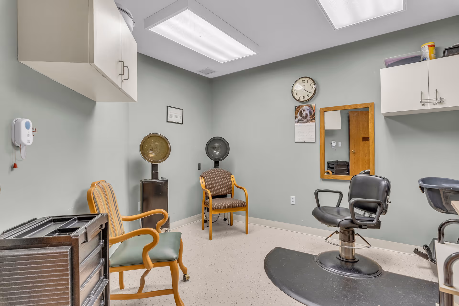 Small salon/hair-styling room with chairs, hooded dryers, a styling chair, mirror, and overhead cabinets.