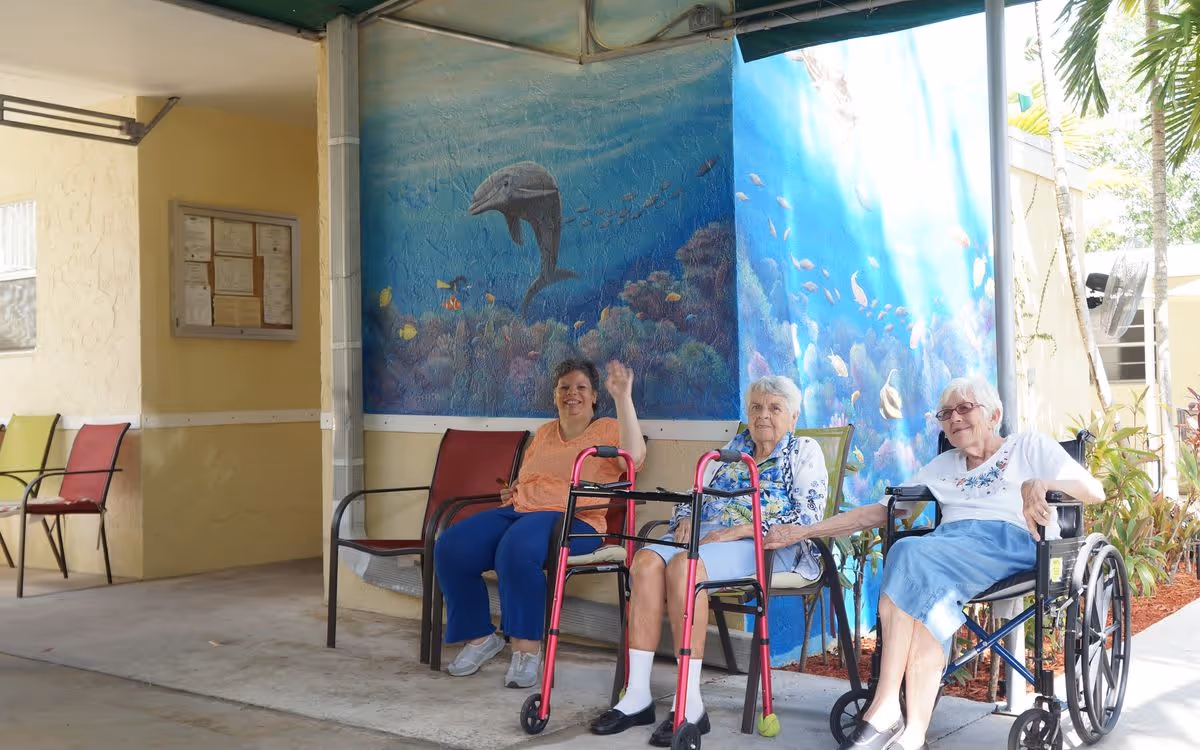 Three elderly women sitting outside near a wall painted with an underwater ocean mural featuring a dolphin and various fish. One woman is in a wheelchair, another is sitting on a chair with a walker in front of her, and the third woman is sitting on a chair waving. The area is shaded and has additional empty chairs nearby.