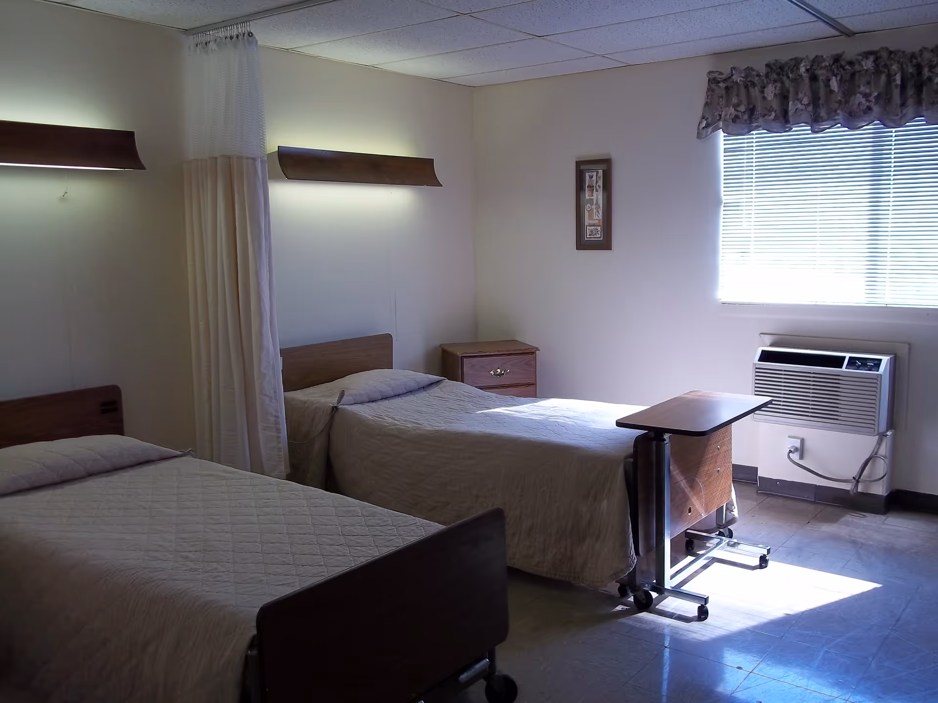 A simple room with two single beds covered in beige quilts, a small wooden nightstand between them, a rolling overbed table in front of one bed, a window with blinds and a floral valance, and an air conditioning unit below the window.