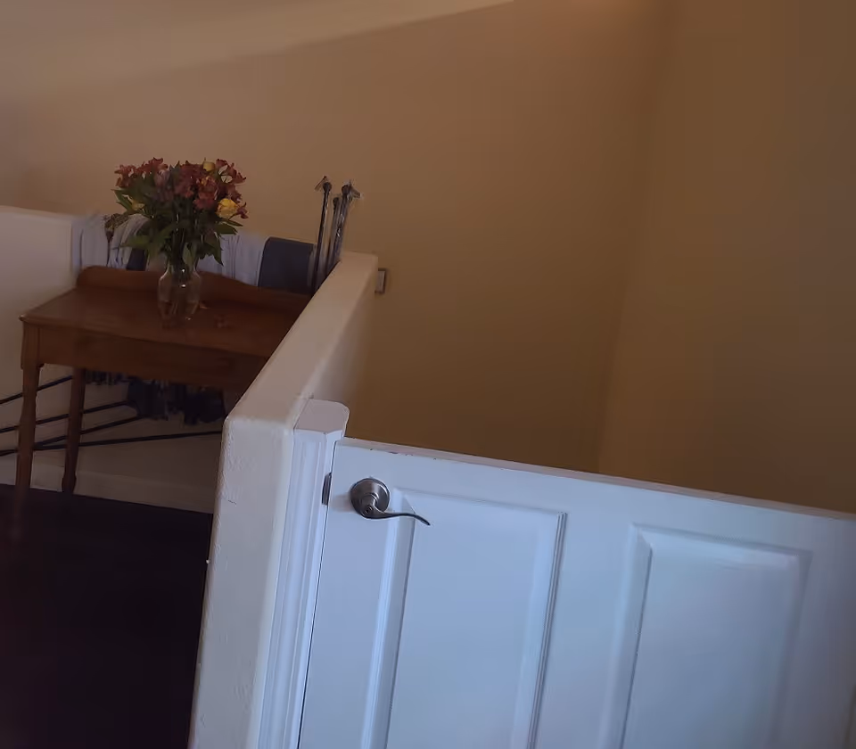 Interior stair landing with a half-door gate, a small wooden table topped with a vase of flowers and walking canes.