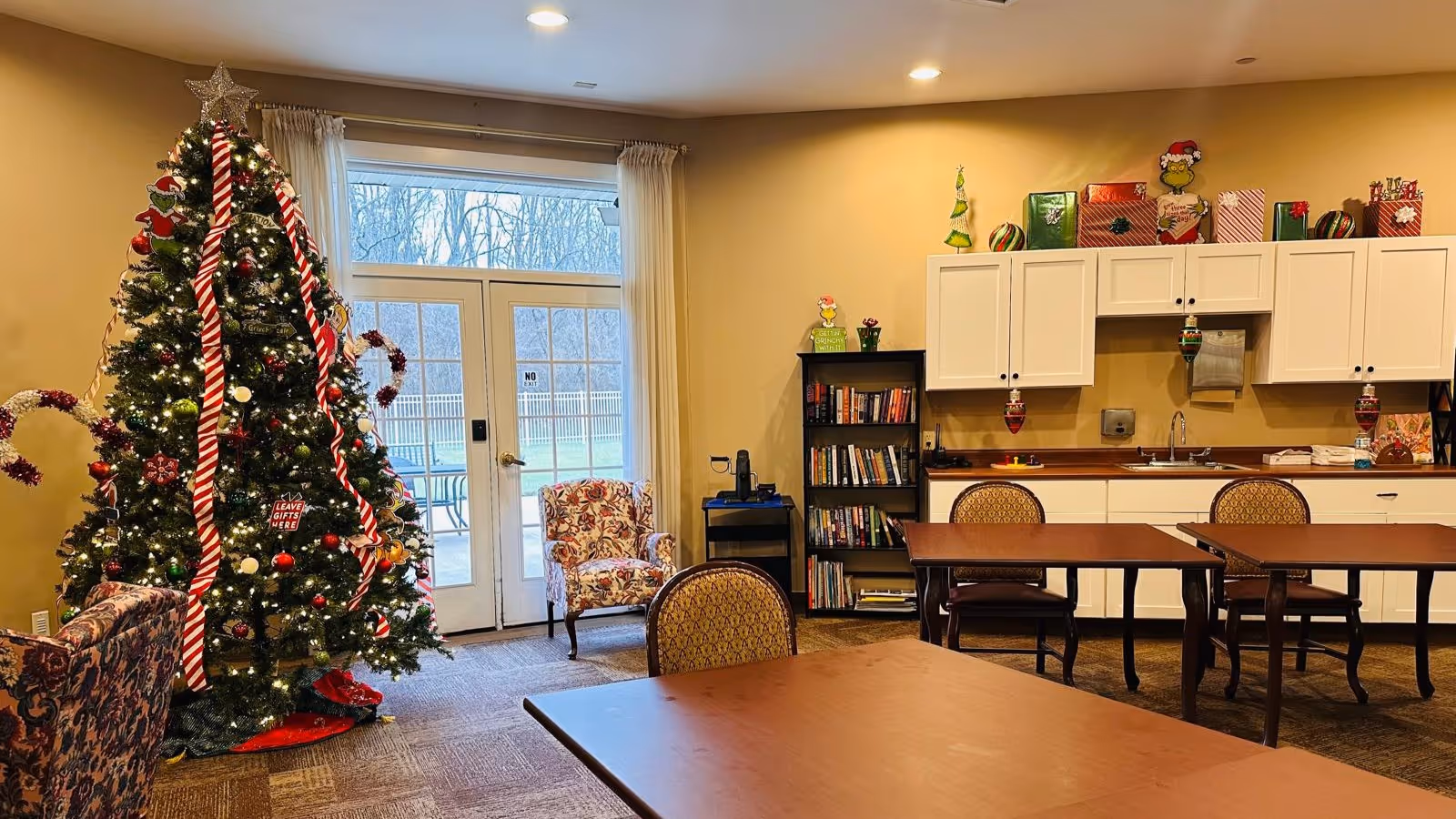 A cozy common area decorated for Christmas with a large Christmas tree adorned with lights, candy cane ribbons, and ornaments. There are several wooden tables and chairs, a floral armchair near glass doors leading outside, a bookshelf filled with books, and white cabinets with holiday decorations on top.