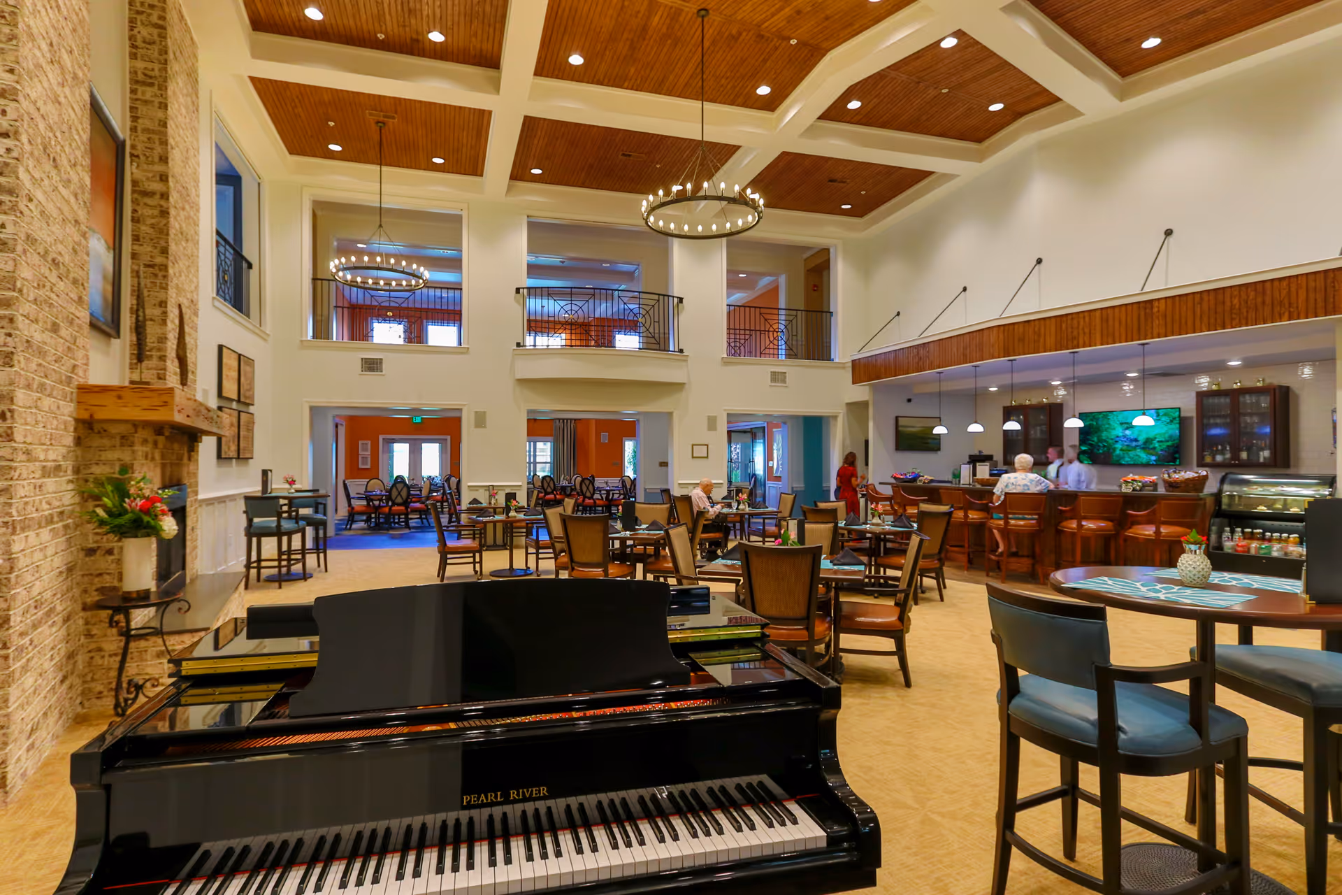 A bright double-height communal lounge and dining area with a grand piano in the foreground, multiple tables and chairs, and a bar under a coffered ceiling.