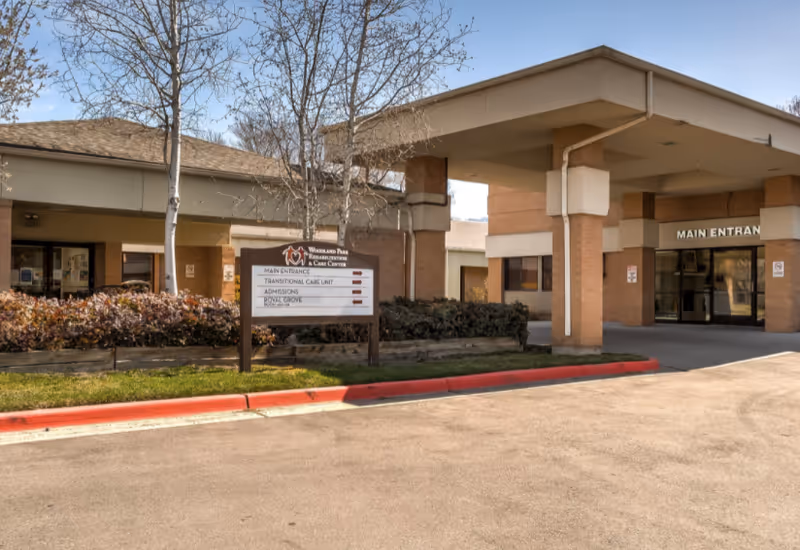 Covered main entrance of a senior living facility with a driveway, canopy, and a directional sign in front.