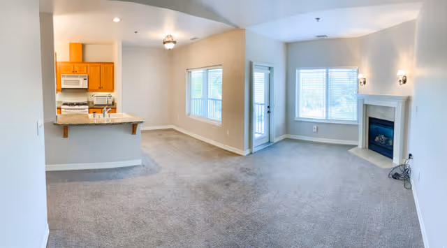 An empty open-concept living space featuring a kitchen area with wooden cabinets, a microwave, and a stove on the left. The kitchen counter has a granite countertop with a sink. Adjacent to the kitchen is a dining area with a window and a ceiling light fixture. The living room area has a large window, a glass door leading outside, and a fireplace with two wall-mounted lights above it. The floor is carpeted throughout, and the walls are painted white.