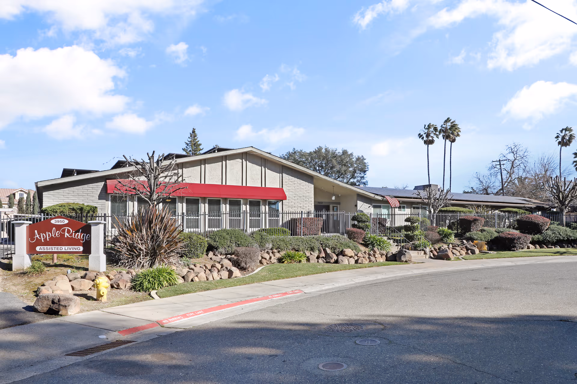 Exterior view of Apple Ridge Assisted Living facility showing a single-story building with a red awning, surrounded by landscaped bushes and rocks, under a partly cloudy blue sky.