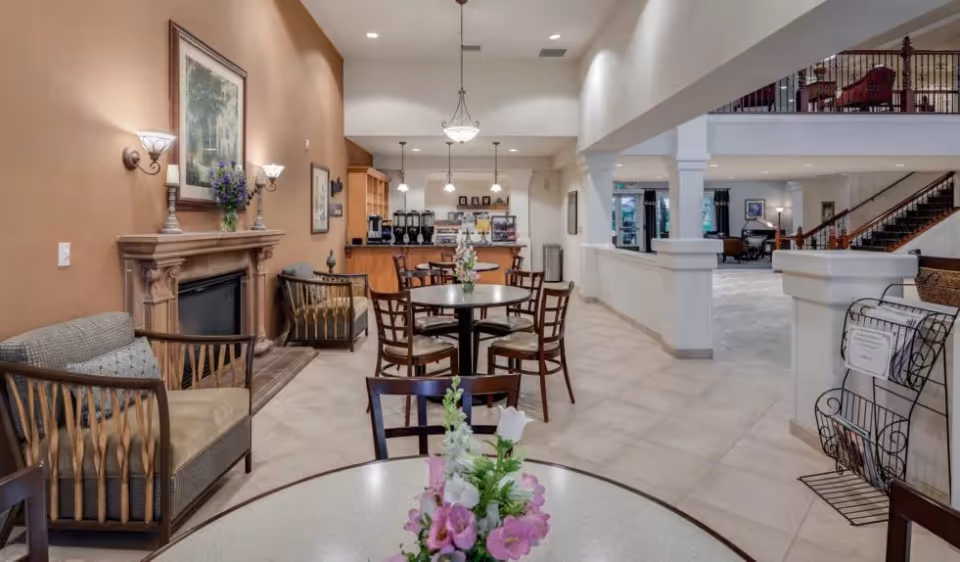 Open communal dining and lounge area with round tables, chairs, a fireplace and a coffee station in a senior living facility.