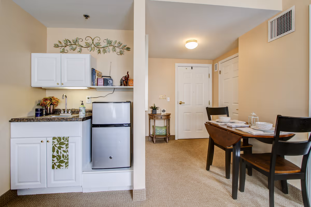 A small kitchenette area with white cabinets, a sink, a mini refrigerator, and a countertop with a few items including a coffee maker and soap dispenser. To the right, there is a wooden dining table set for two with bowls, plates, and a small lantern. The background shows a beige wall with a white door and a small round table with a plant and decorative items. The room has beige carpet flooring and neutral-colored walls.