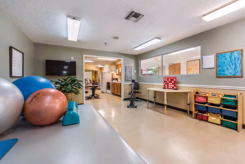 A rehabilitation room in Seneca Health and Rehabilitation Center featuring large exercise balls, a stationary exercise bike, a table with a large Connect Four game, colorful storage bins, and a view into a kitchen area with wooden cabinets and appliances.