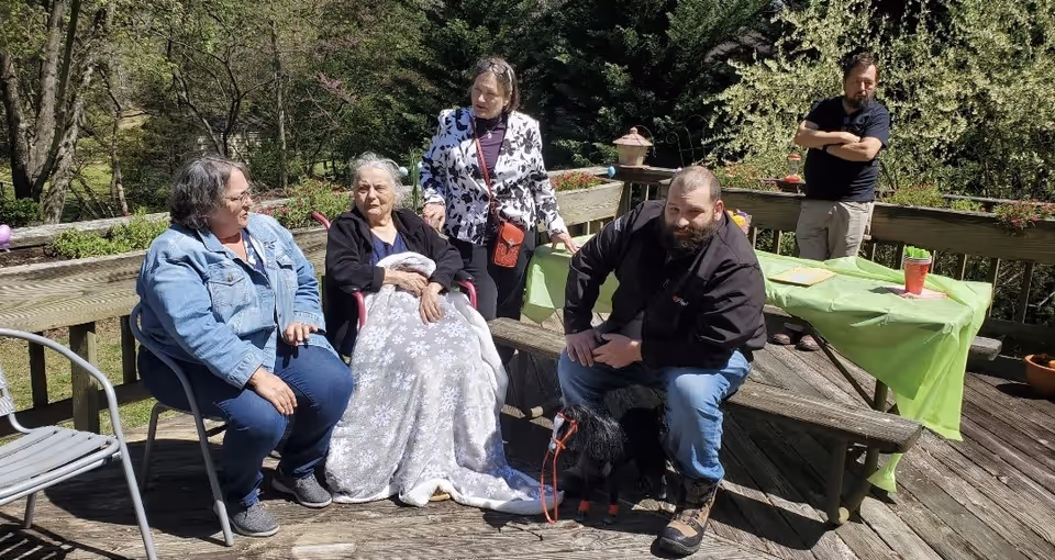 A group of five people gathered outdoors on a wooden deck surrounded by greenery. One elderly woman is seated in a wheelchair covered with a blanket, accompanied by two women and two men. One man is sitting on a bench holding a small black dog on a leash. There is a picnic table with a green tablecloth and some items on it in the background.