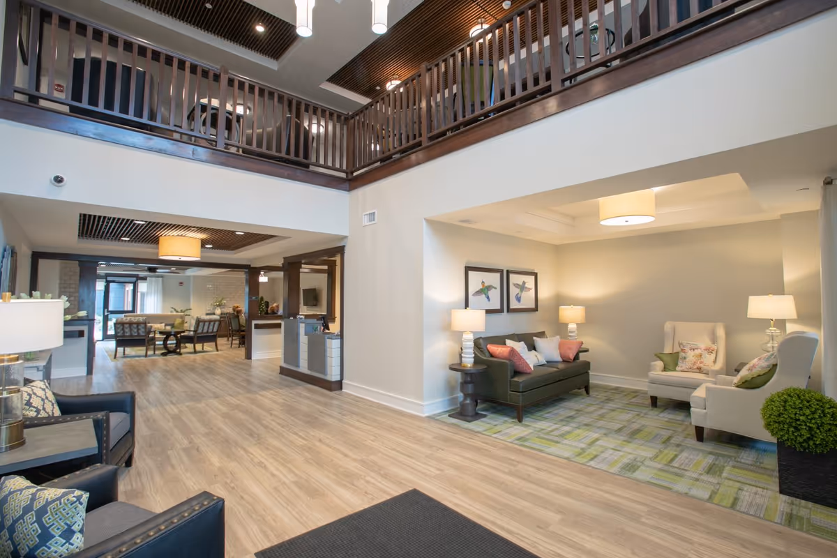 Spacious and well-lit senior living facility common area with wooden flooring, a seating nook featuring a green sofa and two beige armchairs with lamps and framed bird artwork on the wall, and an open view of a lounge area with additional seating and tables. The space has a high ceiling with wooden railings on the upper level and modern light fixtures.
