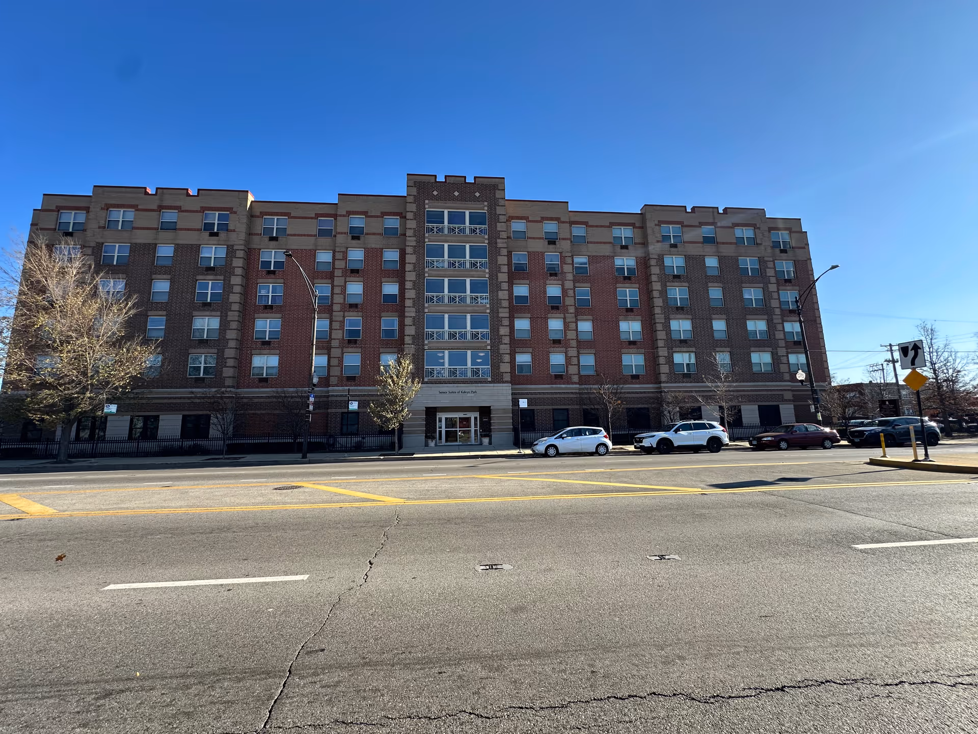 Front view of a six-story brick apartment-style senior living building with parked cars along the street under a clear blue sky.