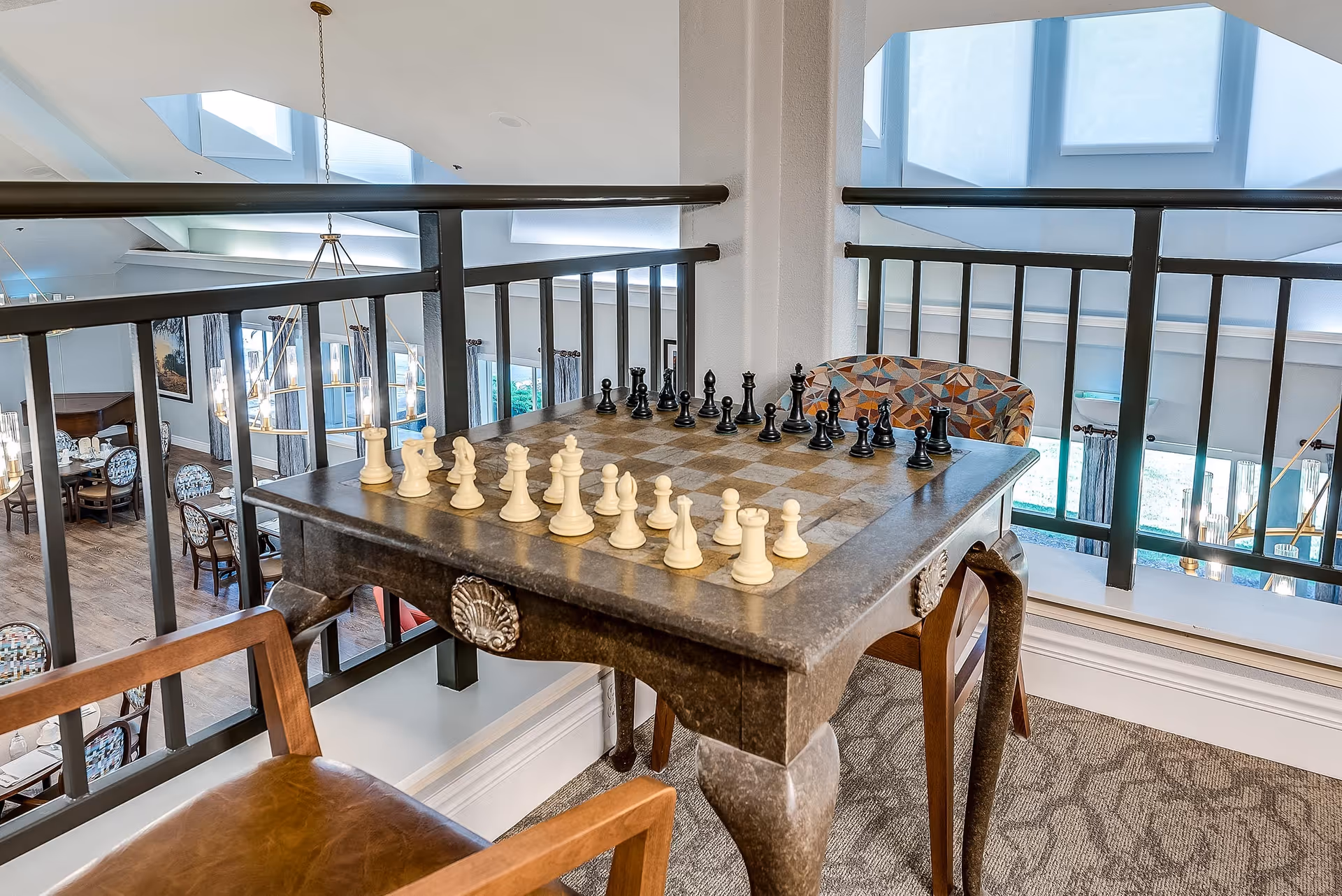 A chessboard set up on a small table by a railing overlooking a dining area in a senior living facility.