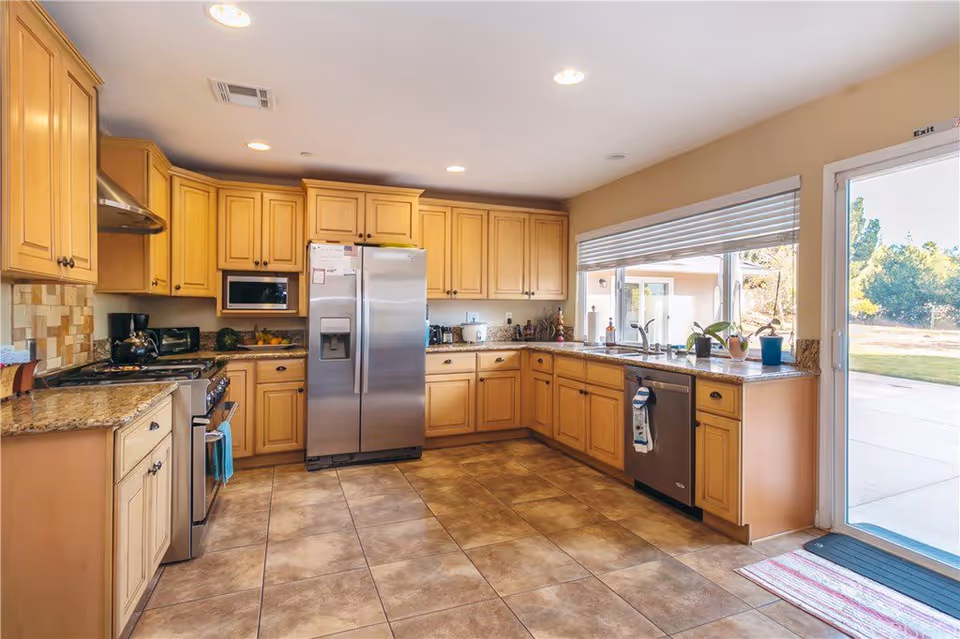 A spacious kitchen with light wood cabinets, granite countertops, and tiled floor. The kitchen features a stainless steel refrigerator, stove, microwave, and dishwasher. There is a large window above the sink with several potted plants on the counter. A glass door leads outside to a patio and grassy area.