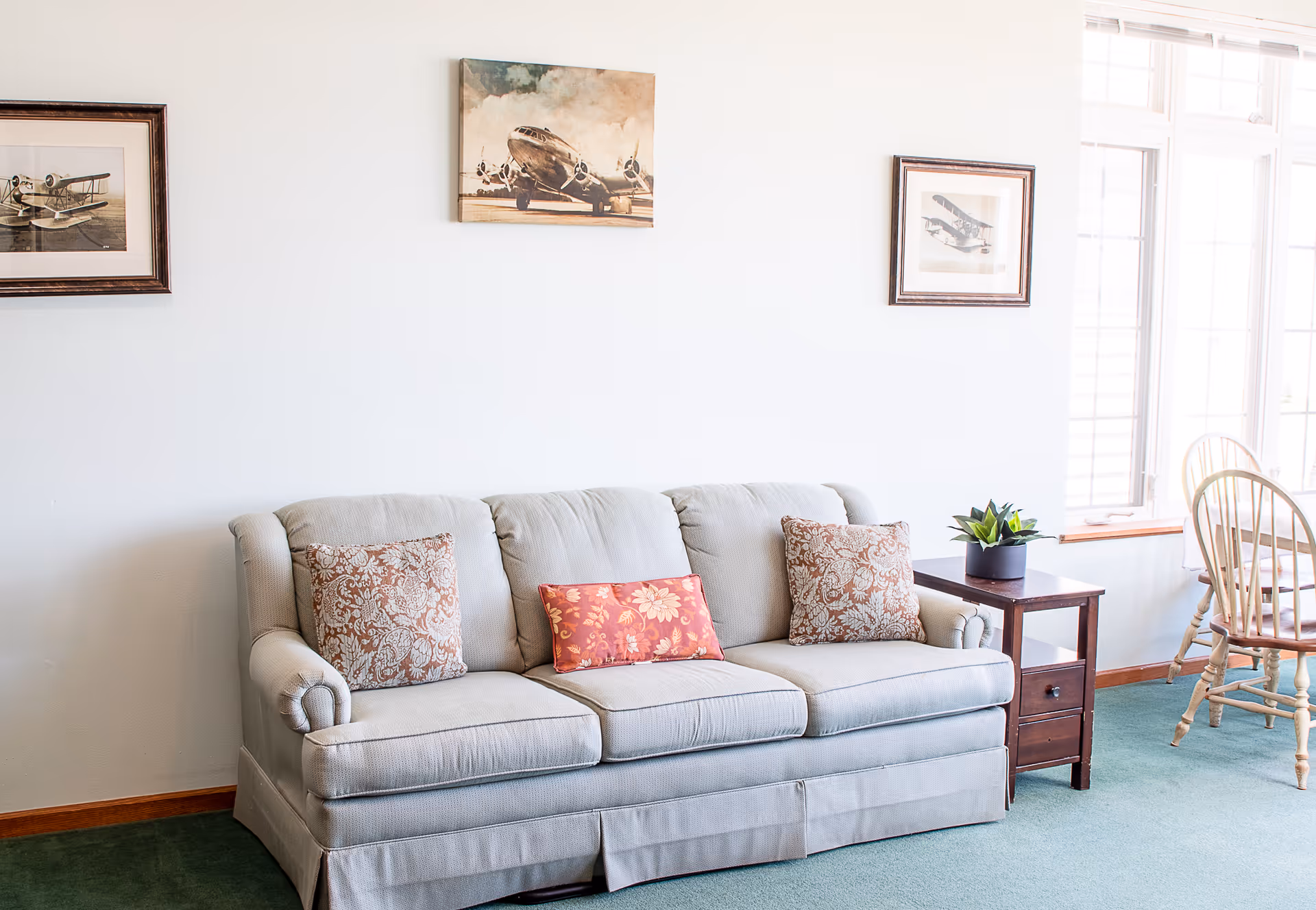 A light gray upholstered sofa with three decorative pillows, two with a floral pattern and one red with a floral design, sits against a white wall. Above the sofa are three framed pictures of vintage airplanes. To the right of the sofa is a wooden side table with a small potted plant. In the background, there is a window with white trim and a wooden chair with a round backrest.