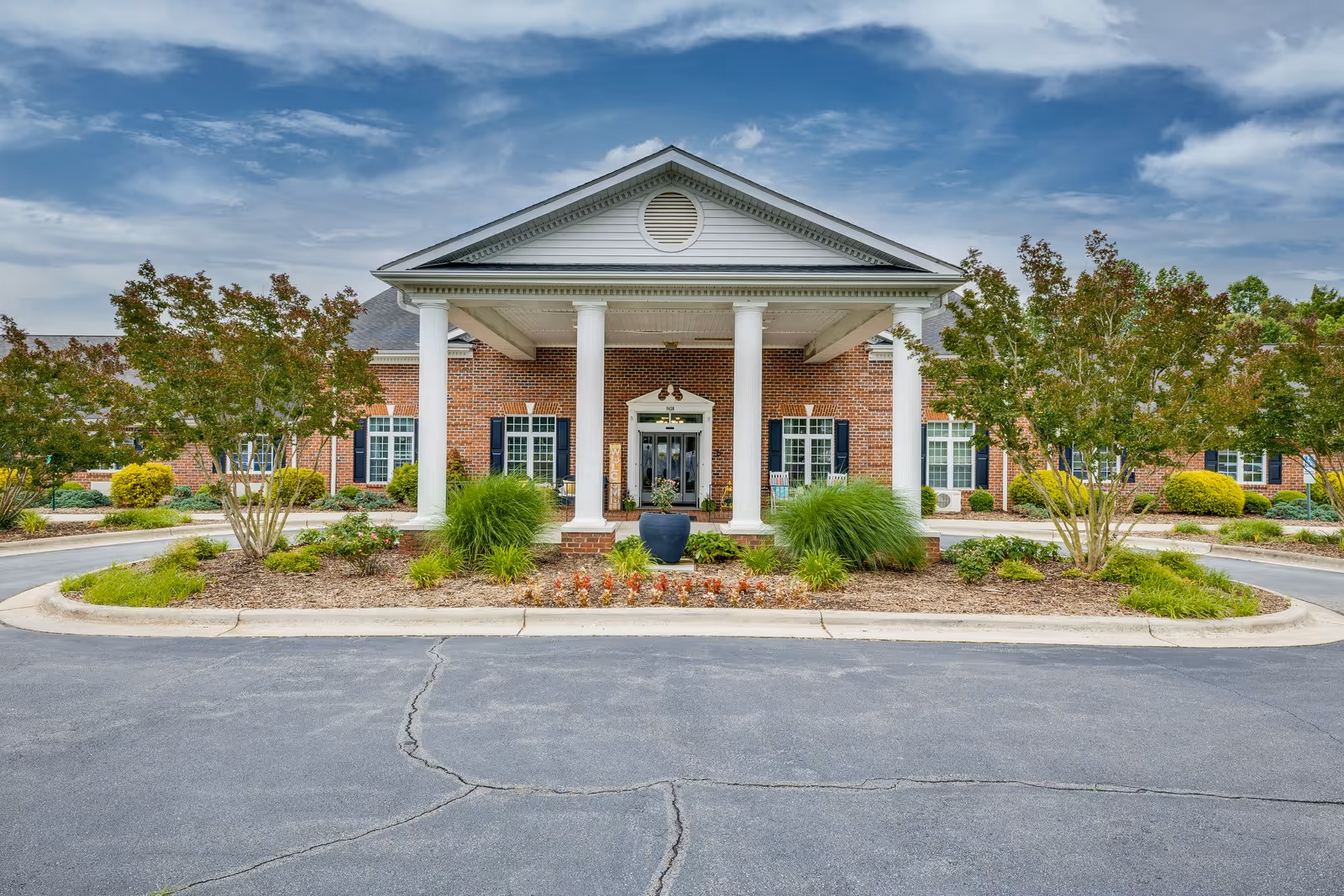 Front exterior view of Mallard Ridge Assisted Living facility featuring a brick building with white columns at the entrance, surrounded by landscaped greenery and trees under a partly cloudy sky.