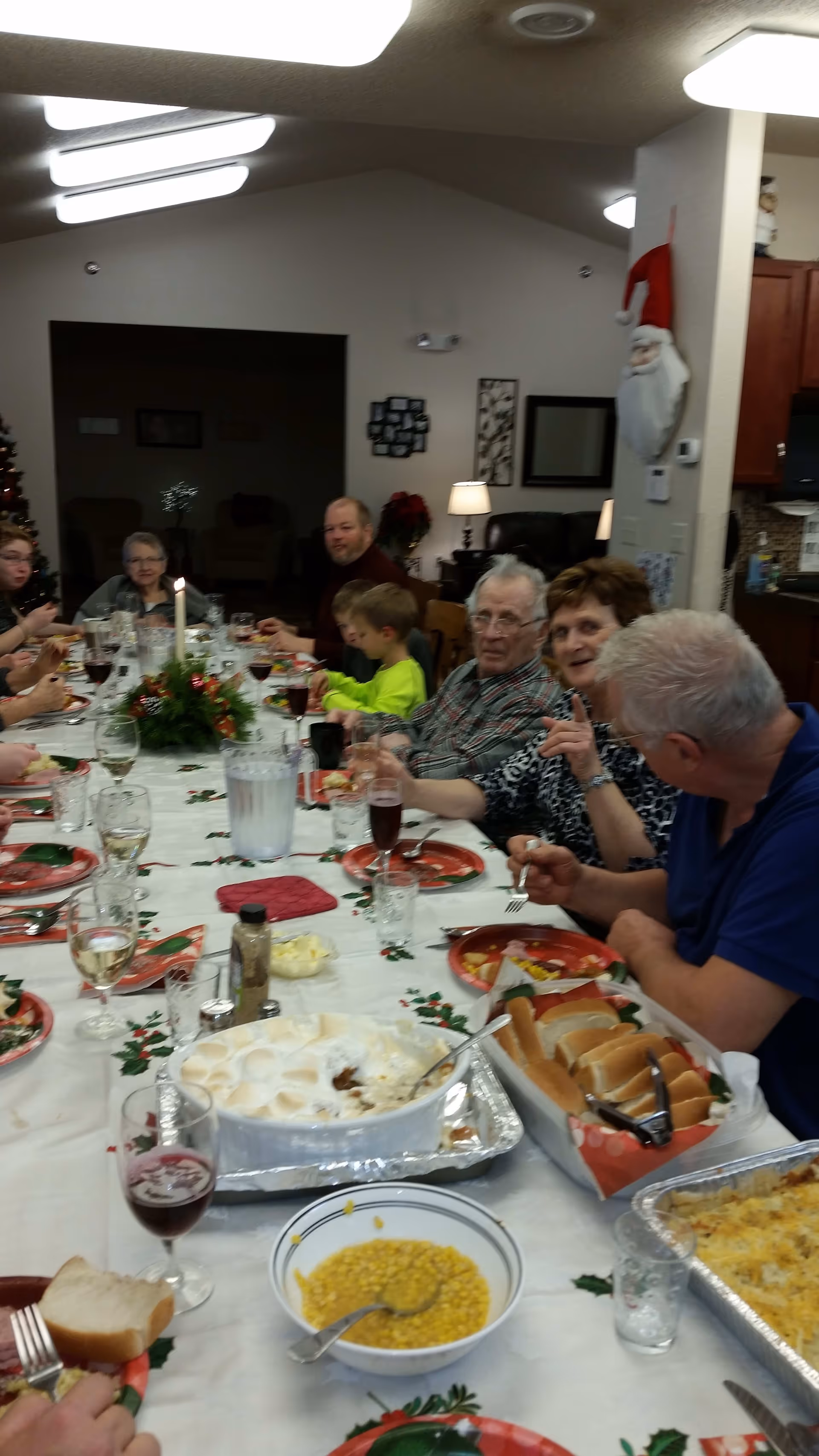 A group of people, including elderly adults and a child, sitting around a long dining table enjoying a meal together. The table is decorated with a white tablecloth featuring holly patterns, a centerpiece with a candle, and various dishes including corn, rolls, and a casserole. The room has warm lighting and festive decorations, including a Santa Claus figure hanging on a pillar.