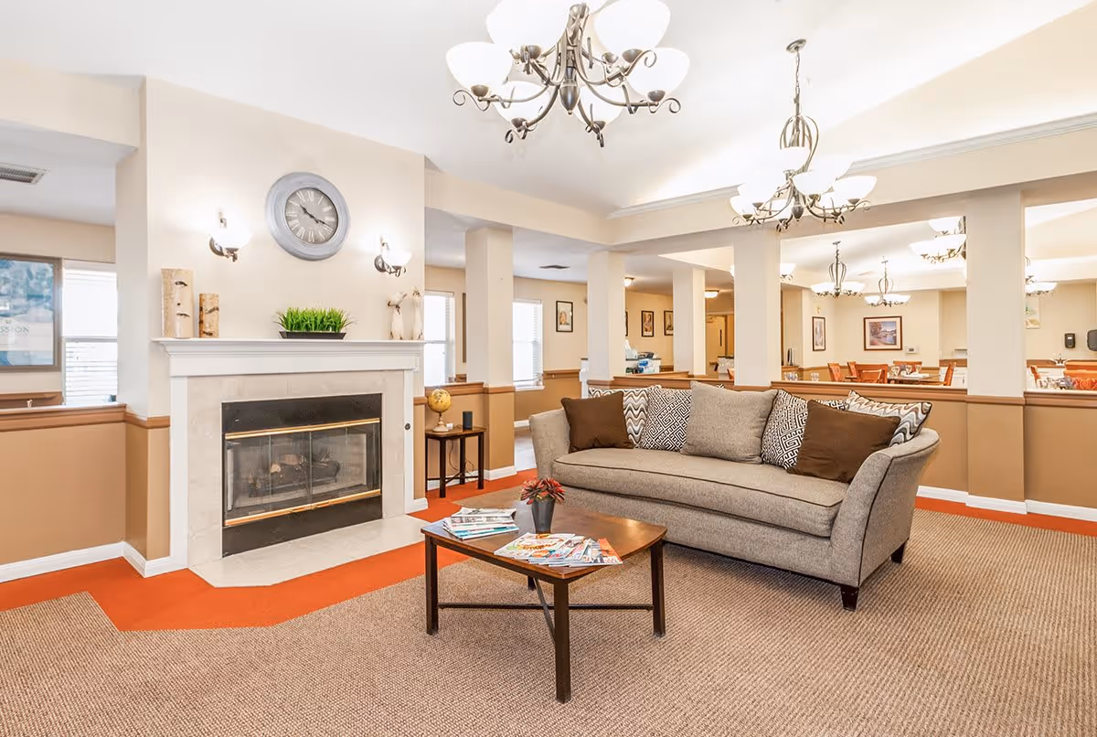 A cozy senior living facility common area featuring a gray sofa with patterned and solid brown pillows, a wooden coffee table with magazines and a small plant, a fireplace with a clock and decorative items above it, and multiple chandeliers hanging from the ceiling. The room has beige walls with brown wainscoting and carpeted flooring with an orange border.
