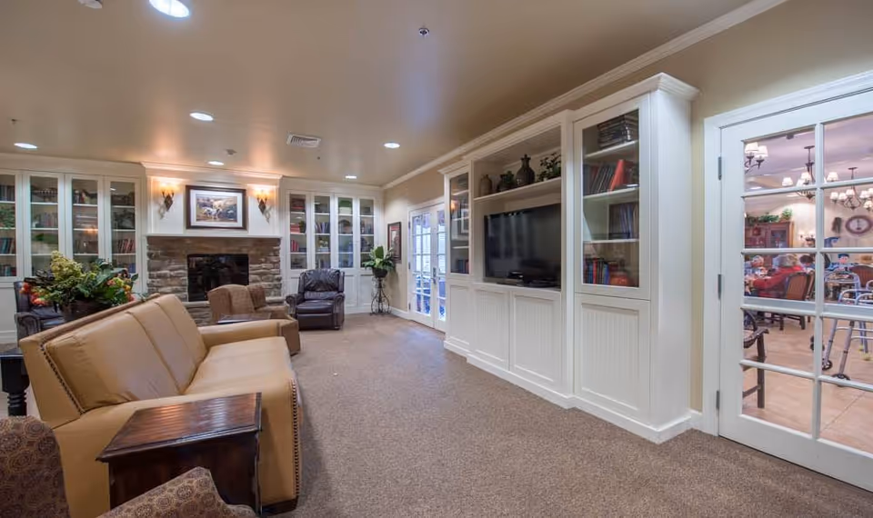A cozy senior living facility common area with a tan leather sofa, armchairs, a stone fireplace, built-in white cabinetry with a TV, and glass doors leading to a dining room where seniors are seated.