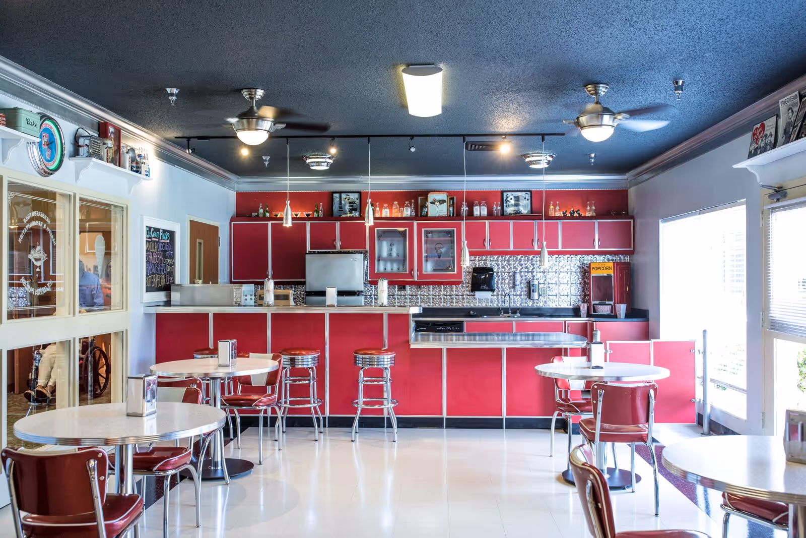 A retro-style dining area with red and white decor featuring round tables with red chairs and a counter with red cabinets and bar stools. The ceiling has fans and pendant lights, and there is a popcorn machine on the right side near a large window.