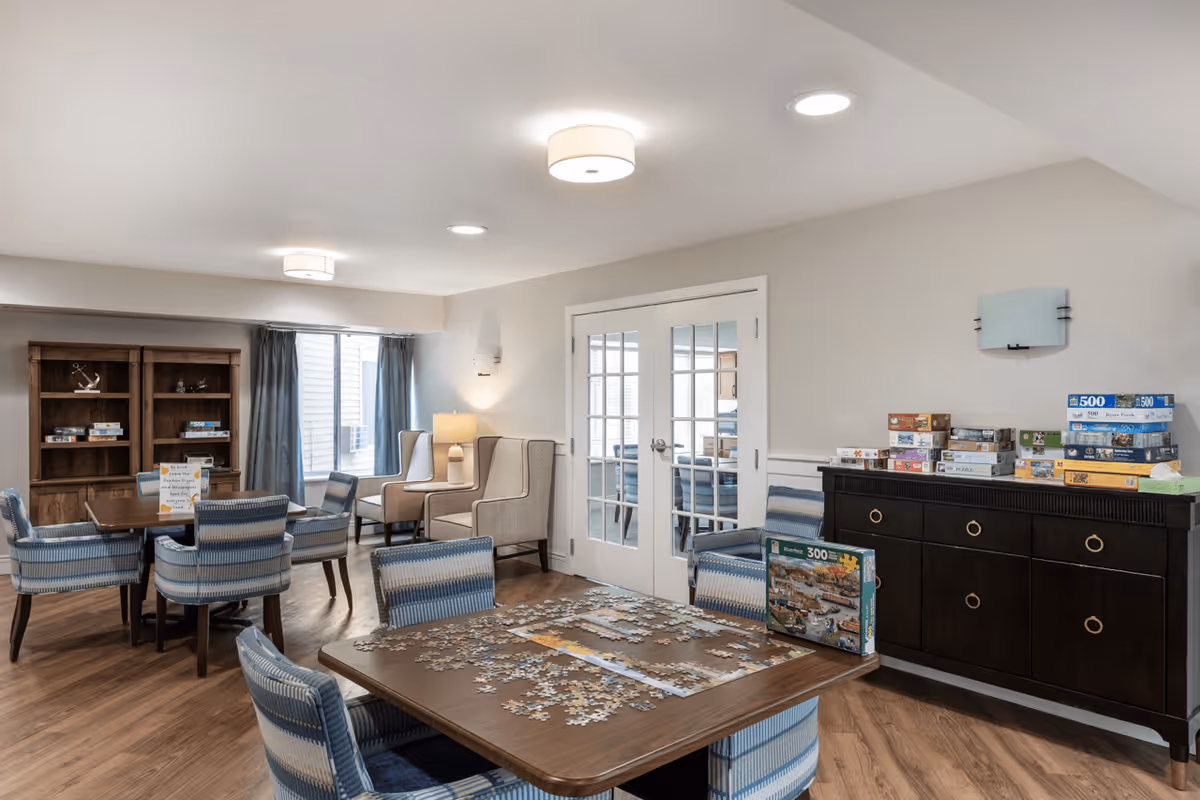 A bright and spacious common area in a senior living facility featuring multiple tables and chairs. One table has a partially completed jigsaw puzzle with puzzle boxes on and around it. There are comfortable armchairs near a window with curtains, a wooden bookshelf with board games, and a dark wooden sideboard with more board games stacked on top. The room has light-colored walls, wood flooring, and ceiling lights.