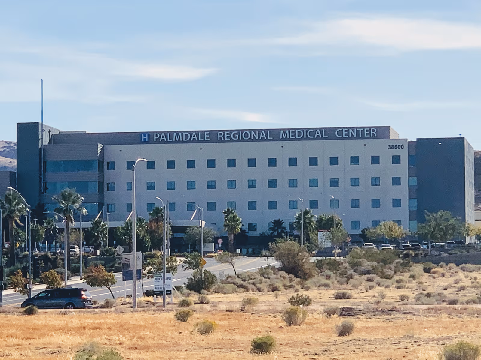 Exterior view of Palmdale Regional Medical Center, a multi-story building with many windows, surrounded by palm trees and a parking area, with dry shrubland in the foreground under a partly cloudy sky.