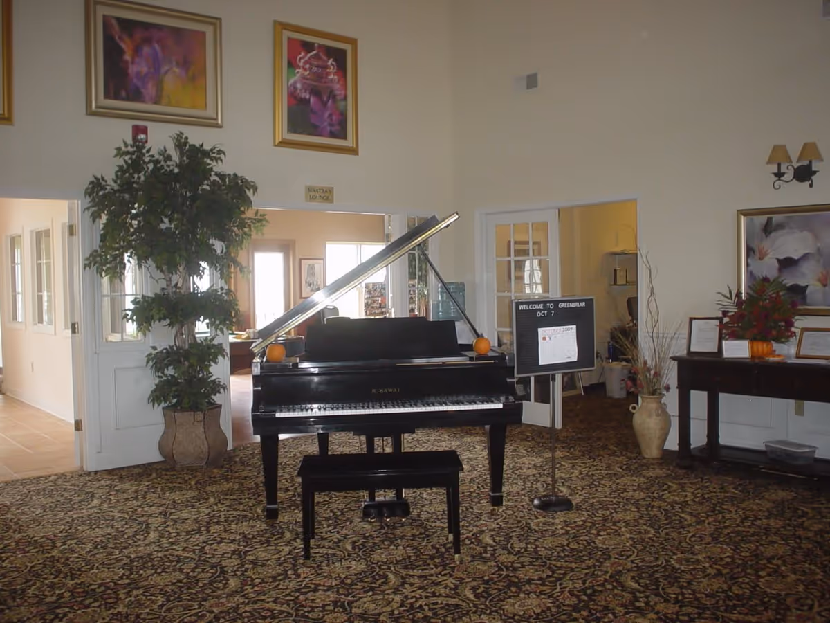 Grand piano in a carpeted common room with a potted plant, welcome sign, and framed artwork on the walls.