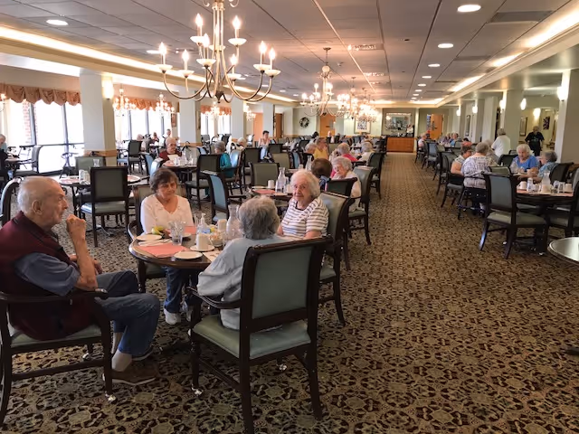 A large dining room in a senior living facility with multiple elderly residents seated at tables, eating and conversing. The room has chandeliers hanging from the ceiling, large windows with curtains letting in natural light, and a patterned carpet floor.