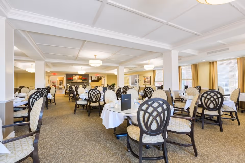 A spacious dining room with multiple round tables covered with white tablecloths, surrounded by cushioned chairs with dark wooden frames and patterned upholstery. The room has beige carpeting, large windows with yellow curtains, and ceiling lights providing warm illumination.