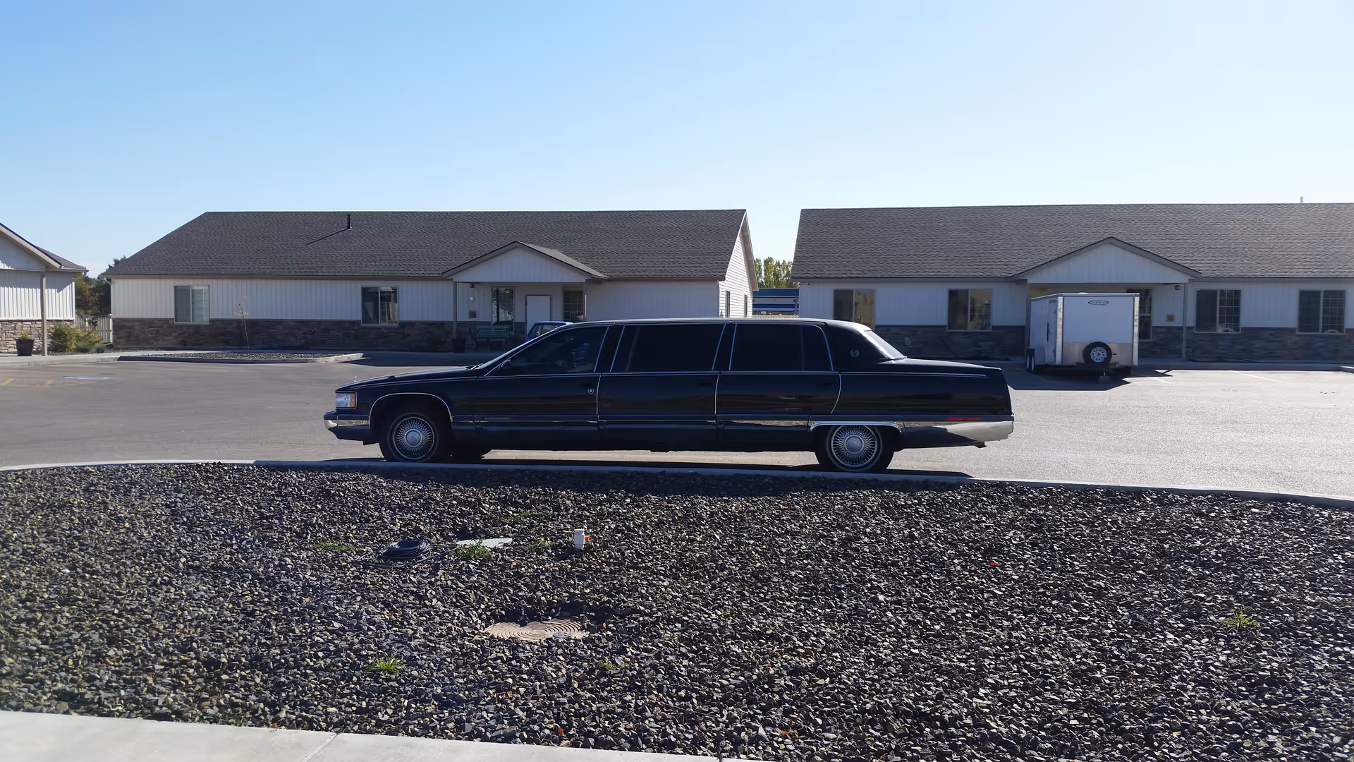 A long black limousine parked on a paved area in front of single-story buildings with white siding and stone accents under a clear blue sky.