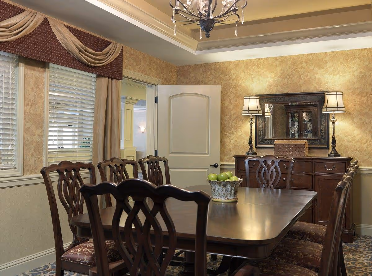 A traditional dining room with a wooden dining table surrounded by eight matching chairs. The table has a decorative bowl filled with green apples. The room features patterned wallpaper, a chandelier hanging from a tray ceiling, and a wooden sideboard with two lamps and a large mirror above it. There are windows with blinds and draped curtains on one side, and a partially open door leading to another room.